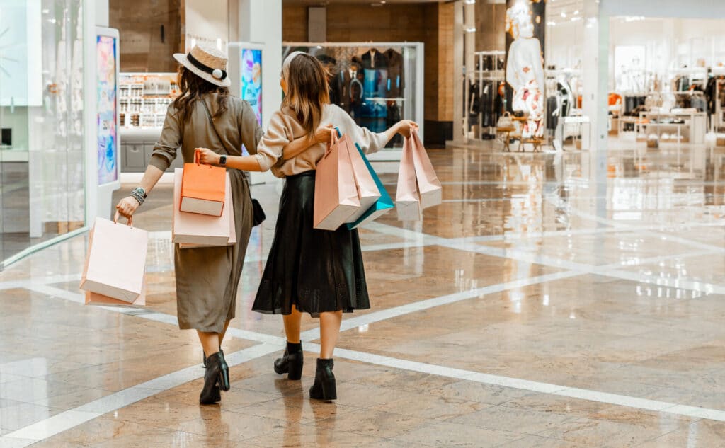 Two shoppers with bags walking through a mall where pop music sets the mood